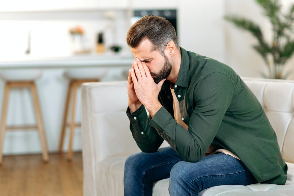 caucasian man dealing with phases of grief, sit on sofa in living room with his head down and his eyes closed